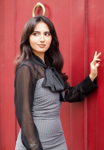 Un vestido gris a rayas con una silueta ajustada, combinado con una blusa negra transparente con un lazo. El fondo es una puerta roja.