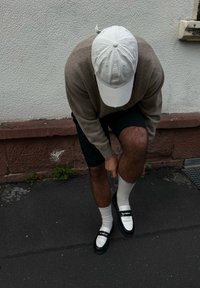 Witte pet, beige trui, zwarte shorts. Draagt witte sokken en zwarte instappers met witte accenten. Staat op een donker asfalt.