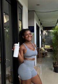 Femme souriante en tenue de sport grise tenant une bouteille d'eau blanche, debout dans un couloir lumineux avec des plantes et un coin salon.