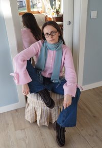 Svetloružový rebrovaný sveter s rozšírenými rukávmi, modrý šál, modré džínsy a čierne topánky. Sedí na pufe so žltými pruhmi. V pozadí drevené detaily.