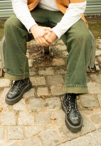 Schwarze Lederstiefel mit Schnürsenkeln und weißen Nähten, getragen mit grünen Hosen, auf einer Kopfsteinpflasterfläche sitzend.