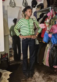 Žalios ir baltos juostelėmis margintas trumpintas kardiganas, tamsios džinsinės šviesos džinsai, mažas rudas krepšys ir žaisminga aplinka su įvairiais drabužiais ir aksesuarais.