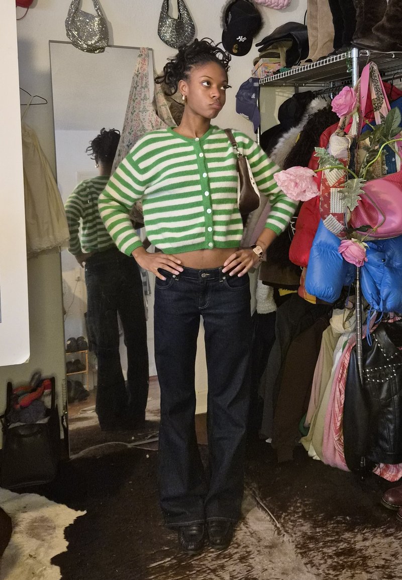 Green and white striped cropped cardigan, dark denim flared jeans, small brown bag, and a playful setting featuring various clothing and accessories.