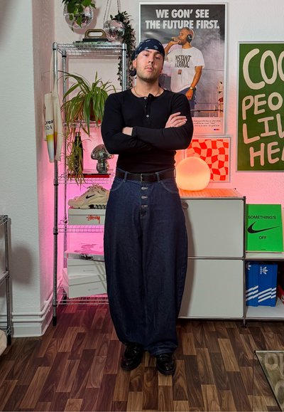 Camisa negra de manga larga, pantalones vaqueros azul oscuro de gran tamaño con grandes botones, zapatos negros y un pañuelo; entorno interior con plantas y decoración.