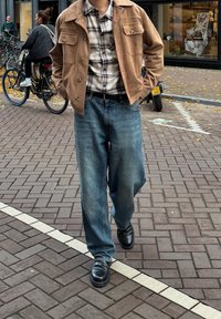 Béžová semišová bunda se dvěma náprsními kapsami, kostkovaná košile, volné modré džíny a černé boty. Městské prostředí s jízdními koly v pozadí.