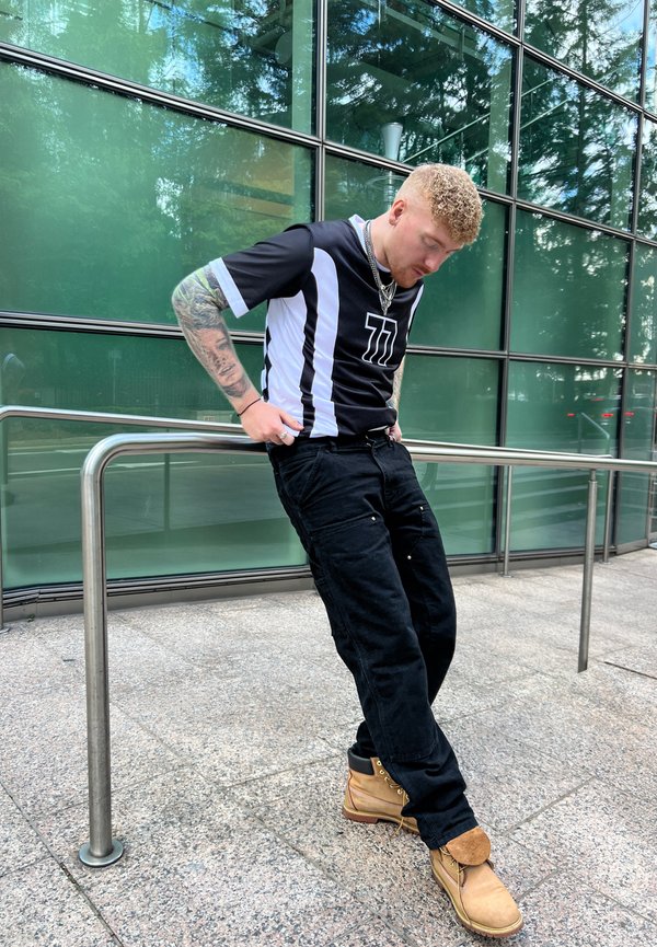 Camiseta atlética en blanco y negro con el número 11, pantalones negros con bolsillos y botas color beige. Enmarcado frente a una pared de vidrio verde.