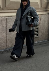 Szürke túlméretezett kabát, alatta fekete kapucnis pulóver, sötét bő szabású nadrág és fekete kézitáska. A megjelenést fekete napszemüveg és sportcipő teszi teljessé.