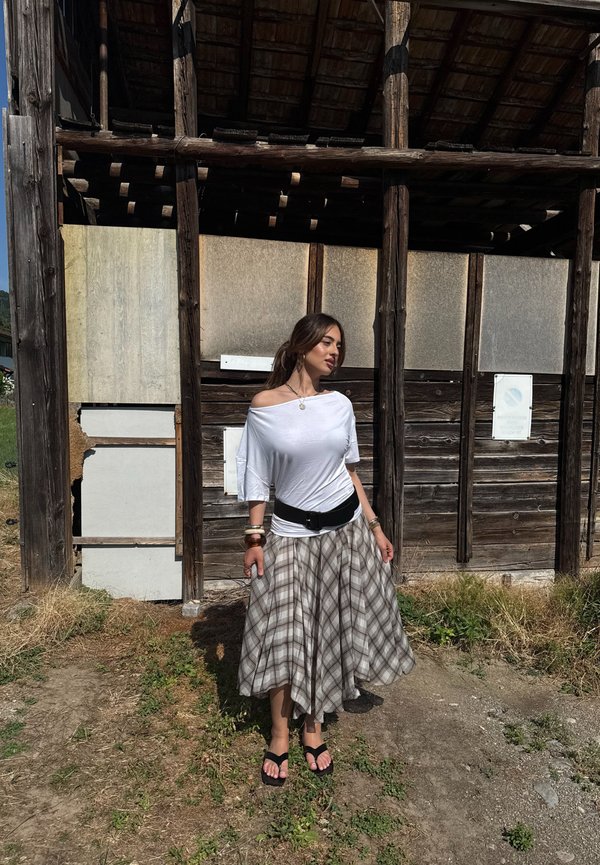 Mujer con un top blanco sin hombros y falda a cuadros, de pie al aire libre junto a una estructura de madera envejecida bajo la luz del sol.