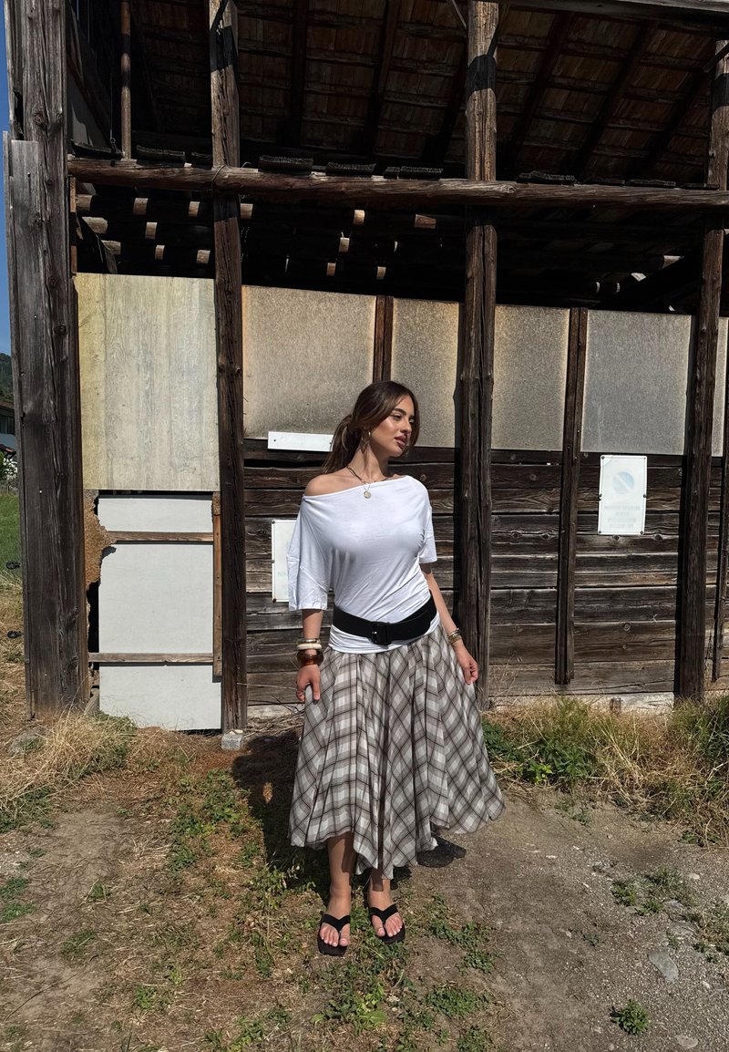 Γυναίκα με λευκό τοπ χωρίς ώμους και καρό φούστα, στέκεται έξω δίπλα σε μια φθαρμένη ξύλινη κατασκευή στο φως του ήλιου.