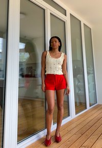 Haut en tricot crème avec des bords froncés, shorts rouges et chaussures rouges. Debout sur un sol en bois près de grandes portes en verre, avec un reflet visible.