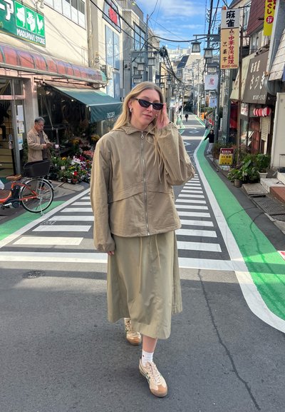 Chaqueta de cremallera beige claro con capucha, combinada con una falda larga beige y zapatillas crema. Paso de peatones a rayas y tiendas en el fondo.