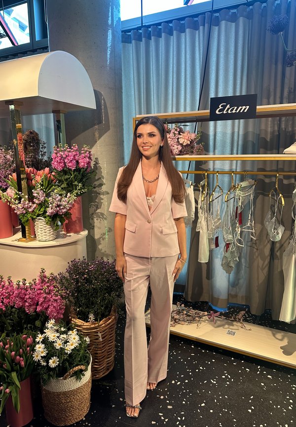 Femeie într-un costum roz pal stă lângă aranjamente florale și un suport de haine cu lenjerie, sub semnul brandului Etam, într-un ambient de boutique.