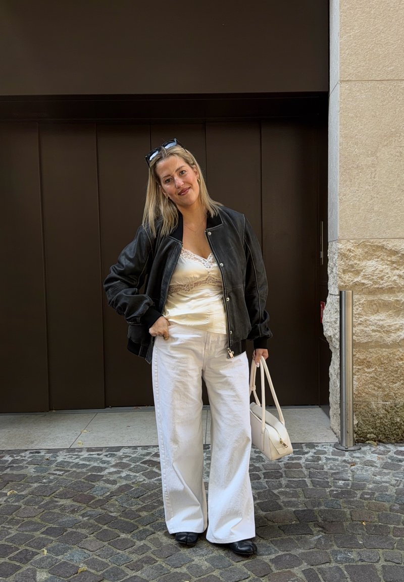 Femme debout dans une rue pavée portant une veste noire, un haut en dentelle blanche, un pantalon blanc large, des chaussures noires et tenant un sac à main blanc.