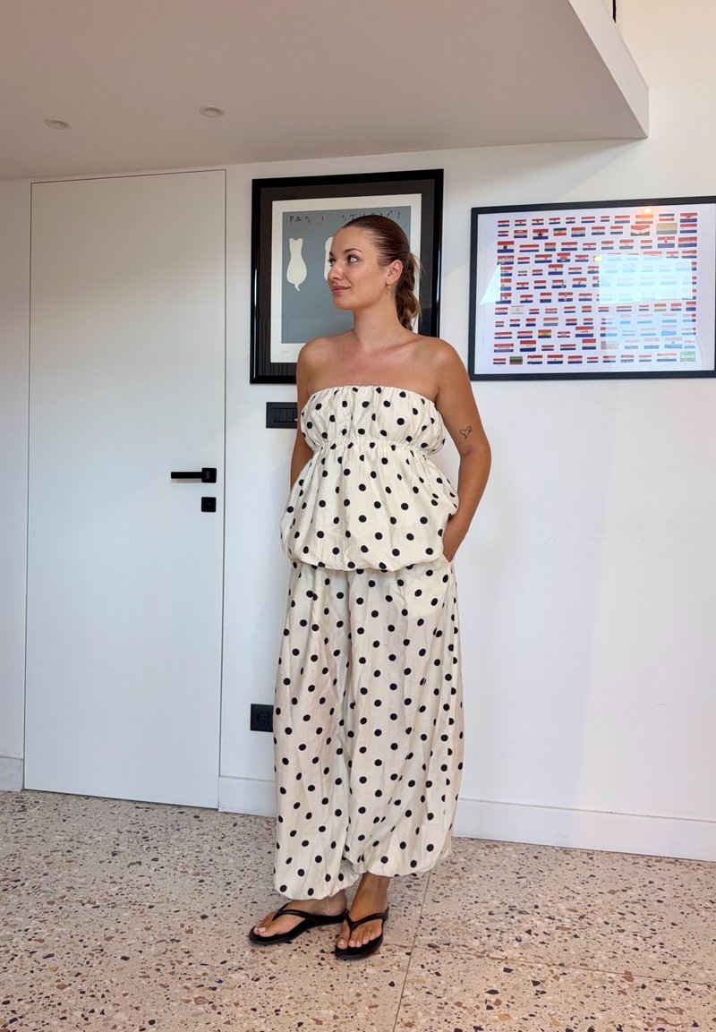 Vrouw in een zwart-wit gestippeld topje zonder bandjes en wijde broek staat blootsvoets op een terrazzo vloer naast een deur en ingelijste wandkunst.