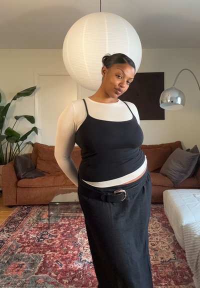 Mujer con top negro sin mangas sobre camisa blanca de manga larga y falda negra está de pie en el salón con un sofá marrón y una gran lámpara colgante blanca.
