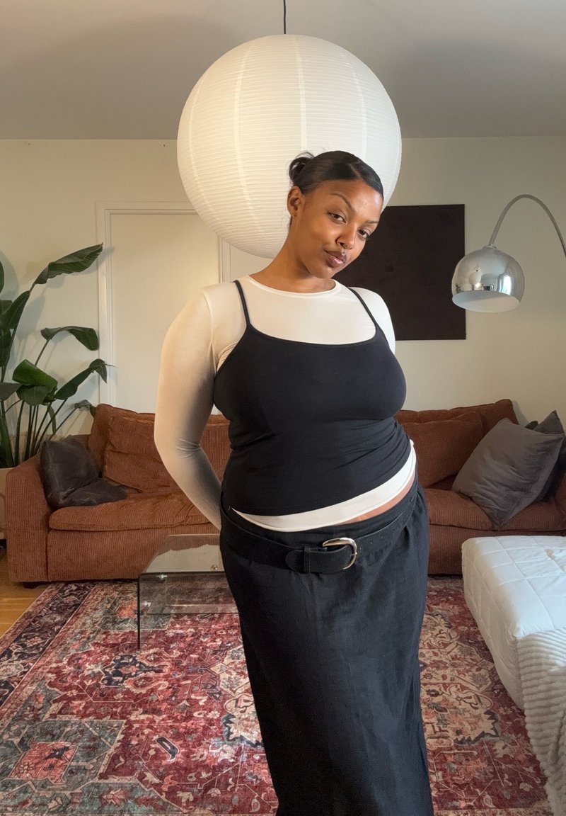 Femme en haut noir sans manches par-dessus une chemise blanche à manches longues et jupe noire, debout dans un salon avec un canapé marron et une grande lampe suspendue blanche.