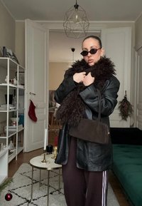 Personne portant une veste en cuir noire, une écharpe sombre et moelleuse, des lunettes de soleil, et un pantalon de survêtement bordeaux tenant un sac à main marron dans un salon moderne.