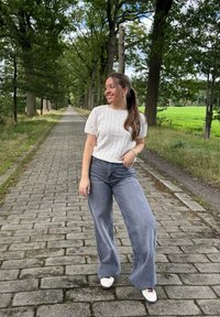 Suéter de malha em bege claro, combinado com jeans cinza de perna larga e tênis brancos. Caminho ladeado de árvores em uma paisagem verde.