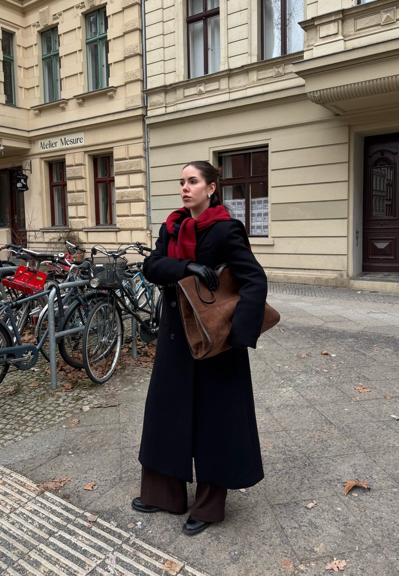 Jeune femme en manteau noir et écharpe rouge se tient près de vélos stationnés devant un bâtiment historique beige.