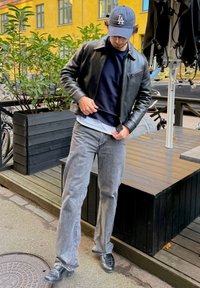 Czarna skórzana kurtka, granatowy sweter, szare dżinsy i czarne buty. Strój jest casualowy; wyróżniające się detale to kieszeń na zamek i warstwowe tkaniny.