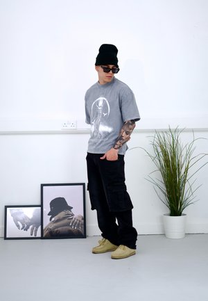 Camiseta gráfica gris con un gran estampado central, pantalones cargo negros y zapatos beige. Los accesorios incluyen gafas de sol y un gorro negro.