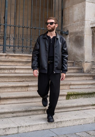 Chaqueta bomber de cuero negra, camiseta polo negra, pantalones holgados negros, zapatos negros con cordones y gafas de sol. De pie en unos escalones de piedra.