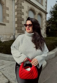 Γκρι ριγέ ζιβάγκο πουλόβερ, κόκκινη τσάντα τύπου κουλούρι, μαύρο παντελόνι, με στρογγυλά γυαλιά ηλίου. Στο ύπαιθρο με φόντο πέτρινο τοίχο και πράσινη βλάστηση.