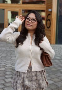Cardigan de punto color beige claro con botones, combinado con una falda de cuadros marrones. Los accesorios incluyen gafas carey y un bolso marrón.