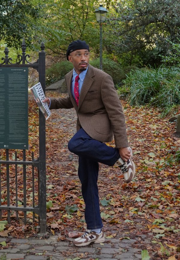 Un homme en veste à carreaux étire une jambe dans un parc verdoyant, un journal à la main, près d’un portail de jardin.