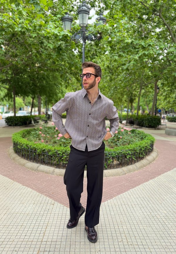 Hombre con gafas que lleva una camisa estampada y pantalones negros, de pie en un camino con baldosas en un parque con vegetación y un poste de luz detrás.
