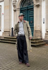 Bărbat îmbrăcat cu o haină verde, un tricou polo gri, pantaloni negri largi, încălțări maronii și o șapcă verde stând pe trotuar lângă o ușă verde și trepte de piatră.