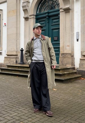 Hombre con abrigo verde, camiseta de polo gris, pantalones anchos negros, zapatos marrones y gorra verde de pie en la acera cerca de una puerta verde y unas escaleras de piedra.
