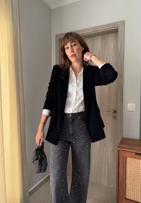 Crni baršunasti sako preko bijele košulje s kopčanjem, upareno s visokim sivim trapericama s bijelim ukrasnim nitnama. Drži torbicu s perlama.