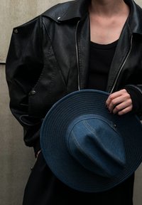 Άτομο που φορά μαύρη δερμάτινη τζάκετ και μαύρο πουκάμισο, κρατώντας ένα ευρύγωνο σακίδιο από μπλε τζην με ραμμένες λεπτομέρειες σε έναν απλό φόντο.