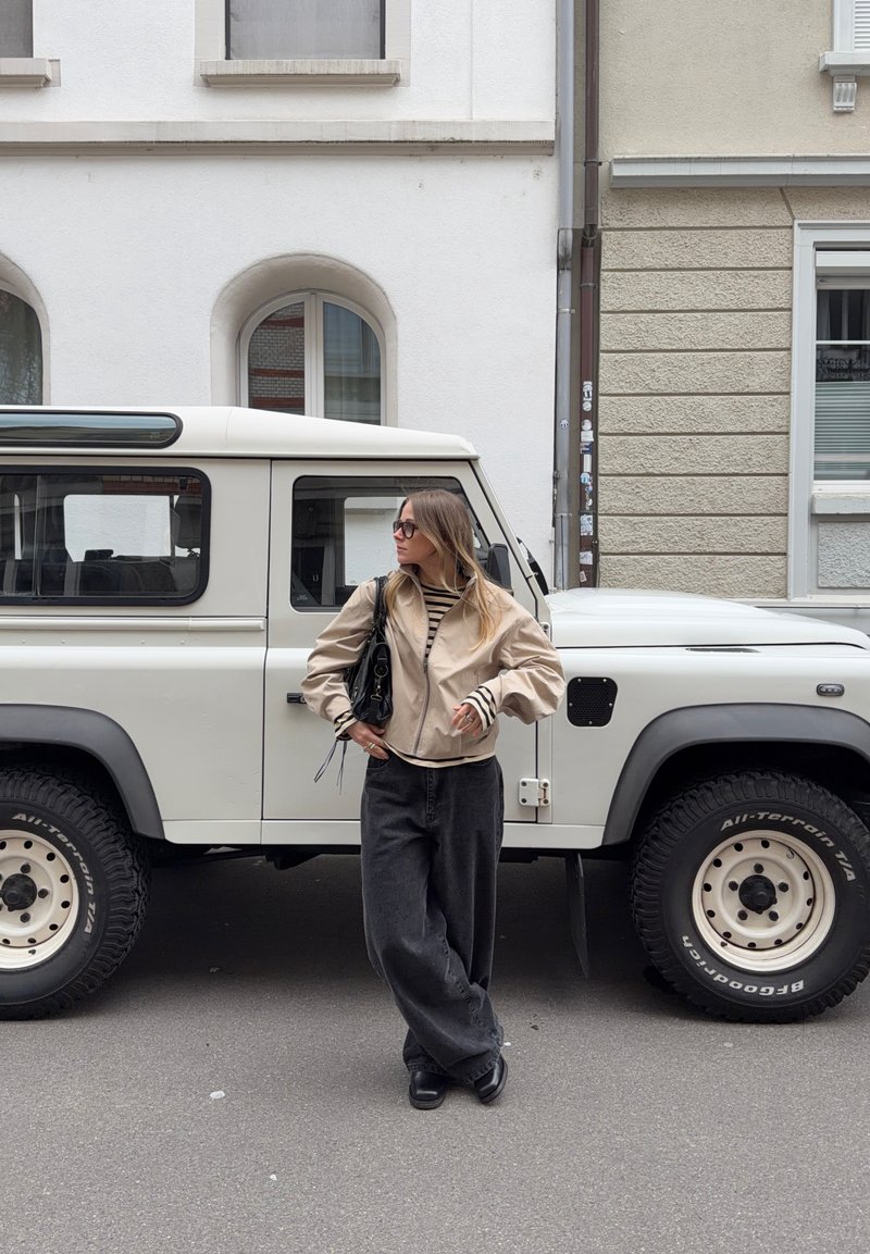 Young woman with long hair wearing glasses and beige jacket stands next to a white off-road vehicle on a city street.