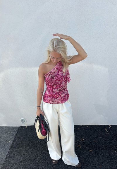 Top asimétrico rosa con un patrón floral, combinado con pantalones de pierna ancha blancos. El cabello es claro y ondulado. Sostiene un bolso negro y de paja.