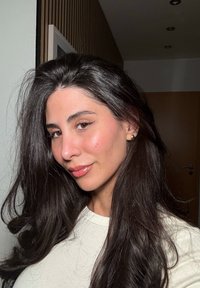 Portret van een persoon met lang, donker, golvend haar, gekleed in een lichtgekleurde shirt en gouden hoepeloorbellen, tegen een zacht verlichte, neutrale achtergrond.