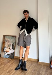 Fekete cipzáros kardigán csíkos póló felett, szürke rövidnadrág és dizájnos aknacipő mellett, minimalista háttér előtt.