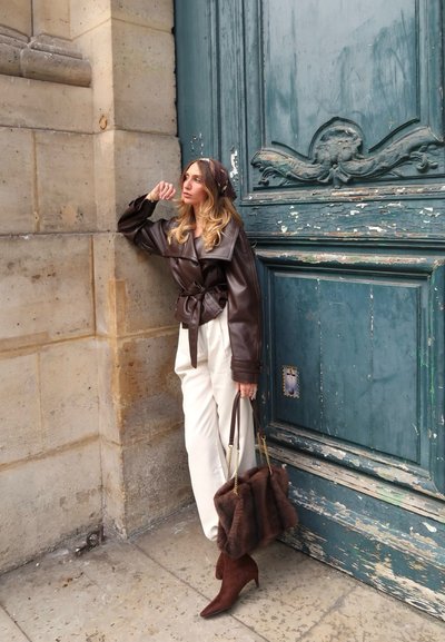 Mujer con chaqueta de cuero marrón y pantalones blancos apoyada contra una puerta de madera azul desgastada, sosteniendo un bolso de piel marrón y llevando botas a juego.