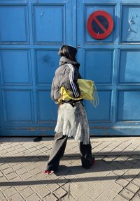 Άτομο με μαύρο μπουφάν και εμπριμέ φούστα περπατά δίπλα σε μπλε ξύλινες πόρτες με πιν ακίδα απαγόρευσης στάθμευσης, κρατώντας μια φωτεινή κίτρινη τσάντα.