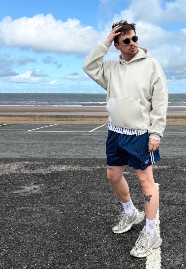 Mand iført solbriller, en cremefarvet hættetrøje, blå shorts og sneakers står på en vej ved havet og rører sit hår med den ene hånd.