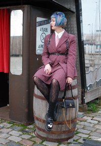 Une personne est assise sur un baril en bois, portant un costume à carreaux bordeaux, une chemise blanche, une cravate rose et des bas en résille noirs avec des chaussures noires épaisses.