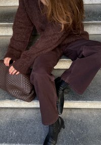 Maglione marrone fluffy, pantaloni a gamba larga marrone scuro, stivaletti neri con tacco e borsa a tracolla intrecciata in marrone, seduta su gradini di pietra.