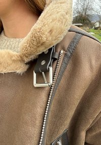 Brun lammeskindsjakke med en kunstpelskrave, sølv lynlås og sort hardware. Blød tekstur med kontrasterende materialer og et klassisk design.