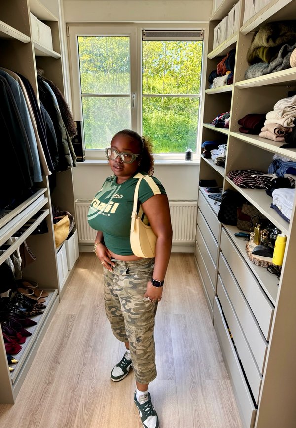 Femme portant des lunettes, un t-shirt vert avec un logo, un pantalon camouflage et des baskets, debout dans un dressing avec des étagères de vêtements pliés et des manteaux suspendus.
