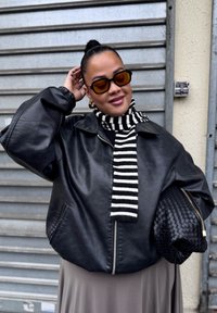 Veste aviateur en cuir noir avec un col montant, écharpe rayée noire et blanche, et un sac à main noir texturé. Lunettes de soleil carrées teintées marron.