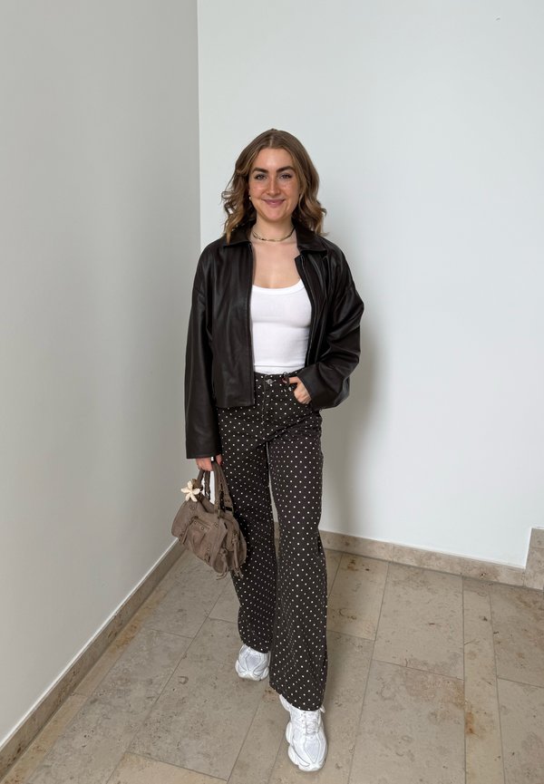 Junge Frau lächelt, trägt schwarze Lederjacke, weißes Top, schwarze gepunktete Hose, weiße Sneaker, hält kleine braune Handtasche drinnen.