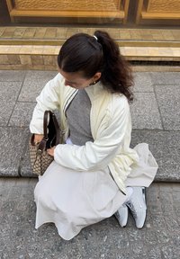 Jachetă albă matlasată, pulover gri tricotat, fustă albă, adidași gri și o geantă cu model. Stând pe piatră cubică, întinzându-se pentru a lua lucruri din geantă.