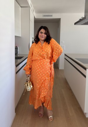 Vestido envolvente naranja con lunares, volantes, mangas largas y detalles de botones. Combinado con un bolso dorado de lentejuelas y tacones con tiras.