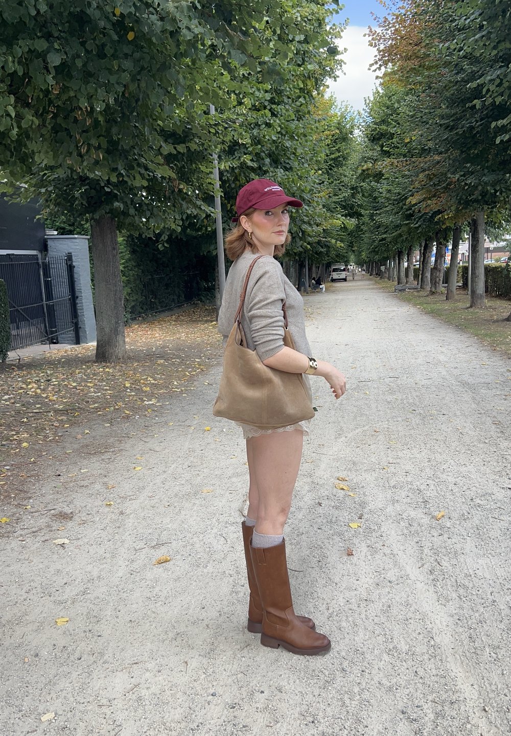 Smeđe kožne čizme do koljena, krem pulover, smeđa torba preko ramena, bordo cap; stoji na šljunčanom putu obrubljenom drvećem.